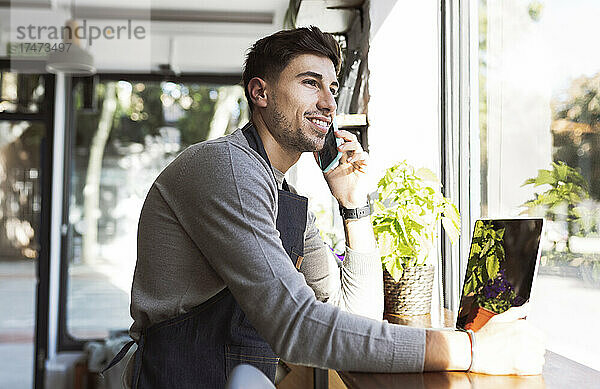 Lächelnder Geschäftsmann mit Laptop  der im Café mit dem Handy telefoniert