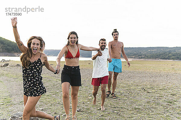 Fröhliche männliche und weibliche Freunde halten sich beim Spaziergang am Seeufer an den Händen