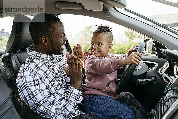 Mann klatscht in die Hände  damit sein Sohn im Auto mit dem Lenkrad spielt