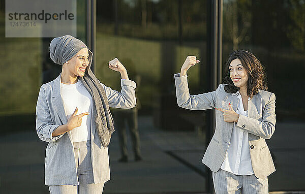 Lächelnde Berufstätige zeigen  während sie ihre Muskeln vor einer Glaswand spielen lassen