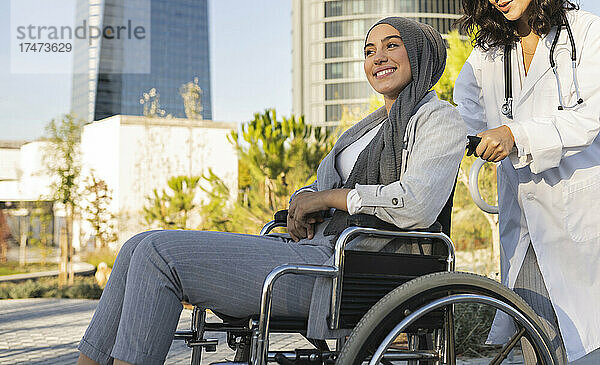 Junge Allgemeinärztin schiebt den Rollstuhl einer lächelnden behinderten Frau auf dem Fußweg