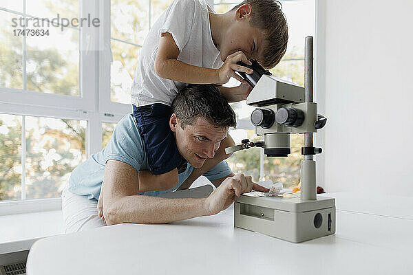 Neugieriger Junge benutzt Mikroskop  während er zu Hause auf den Schultern seines Vaters sitzt