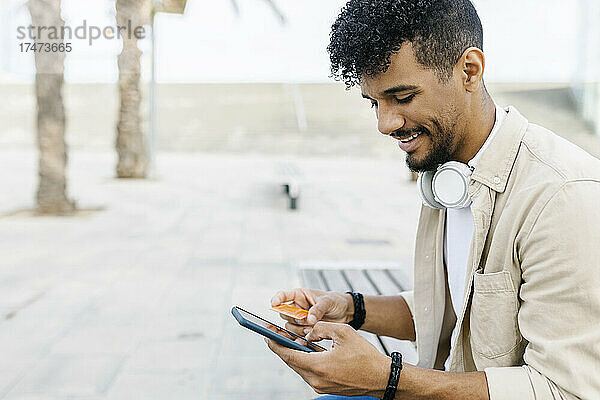 Lächelnder Mann hält Kreditkarte in der Hand und zahlt  während er sein Smartphone benutzt