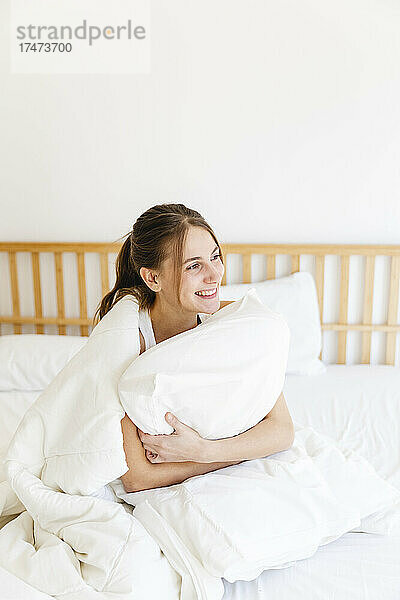 Lächelnde junge Frau umarmt Kissen  während sie im Schlafzimmer auf dem Bett sitzt