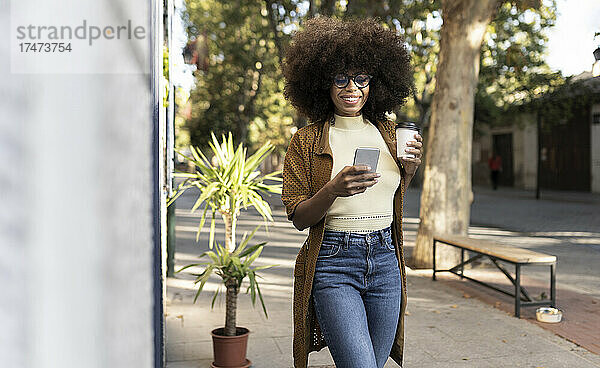 Lächelnde Frau mit Einwegkaffeetasse und Mobiltelefon auf Fußweg