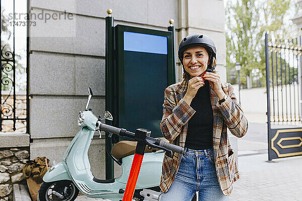 Lächelnde Geschäftsfrau mit Elektroroller und Helm vor dem Park