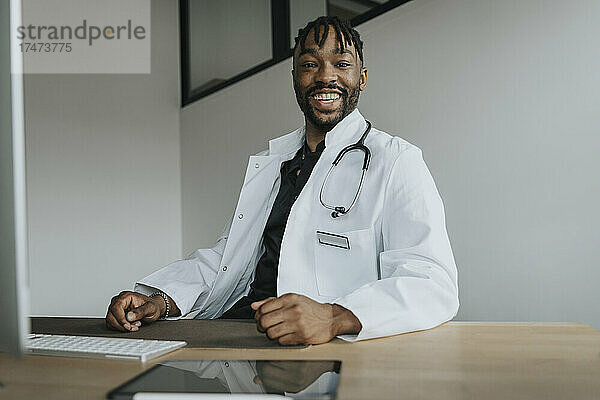 Lächelnder junger Arzt mit Stethoskop am Schreibtisch