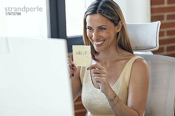 Lächelnde Freiberuflerin zeigt während einer Videokonferenz am Computer zu Hause einen Klebezettel