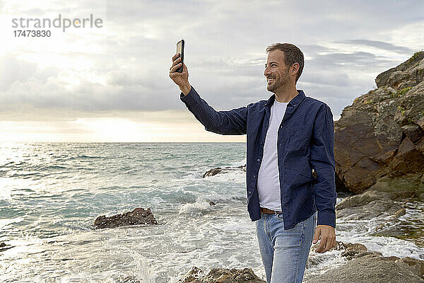Lächelnder Mann  der am Strand ein Selfie mit dem Smartphone macht