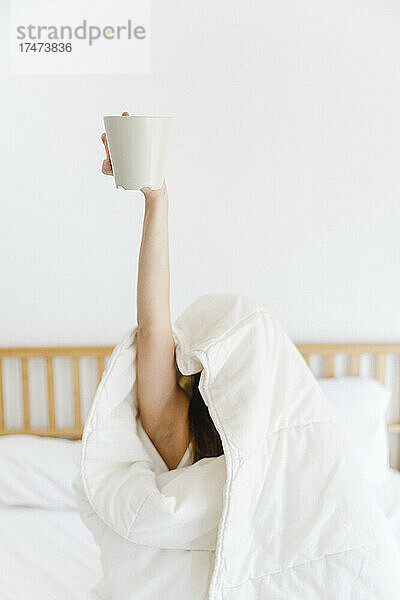 Frau mit erhobener Hand hält Kaffeetasse  während sie in eine weiße Decke gehüllt auf dem Bett liegt