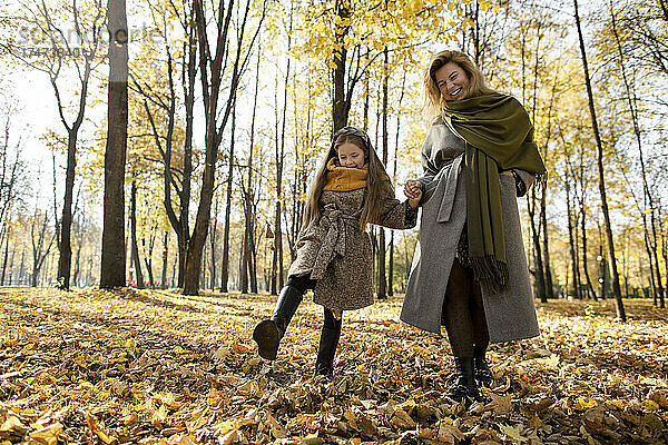 Lächelnde Frau hält die Hand ihrer Tochter und tritt im Park gegen Herbstblätter