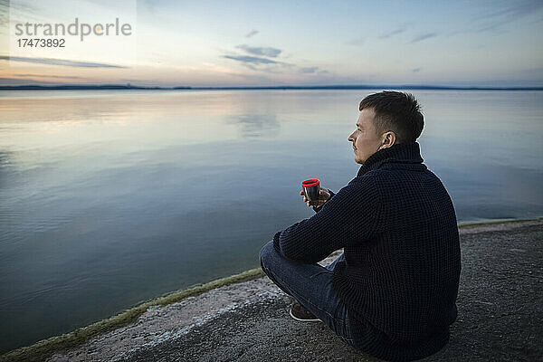Mann trinkt Tee und sitzt bei Sonnenuntergang auf einem Felsen am See