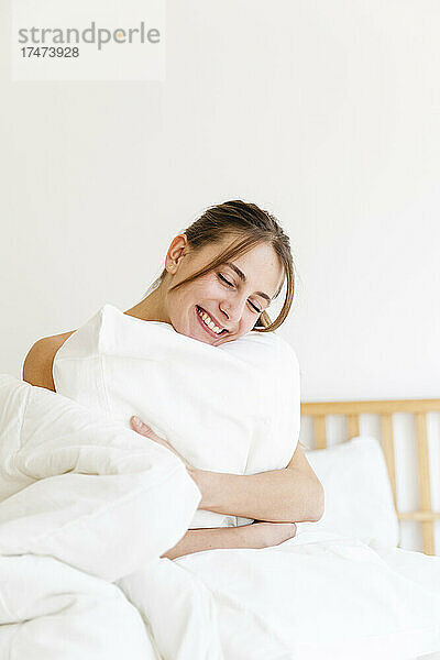 Glückliche junge Frau mit geschlossenen Augen  die zu Hause ein weißes Kissen umarmt