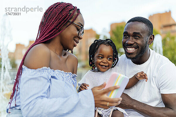 Lächelnder Mann mit Tochter blickt Frau an  die ihr Smartphone zeigt