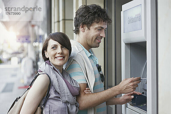 Lächelnder Mann  der neben seiner Freundin steht und Geld vom Geldautomaten abhebt