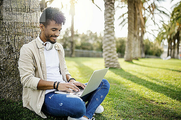 Lächelnder junger Mann benutzt Laptop  während er am Baumstamm sitzt