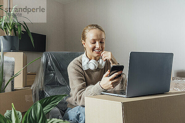 Lächelnde Frau mit Laptop auf Karton und Mobiltelefon in umgezogenem Zuhause