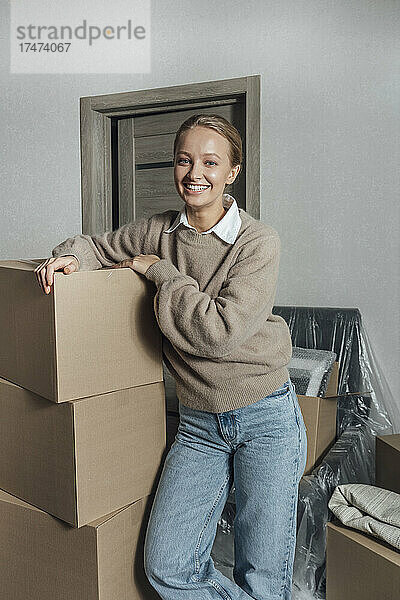 Lächelnde junge Frau stützt sich zu Hause auf einen Stapel Kartons