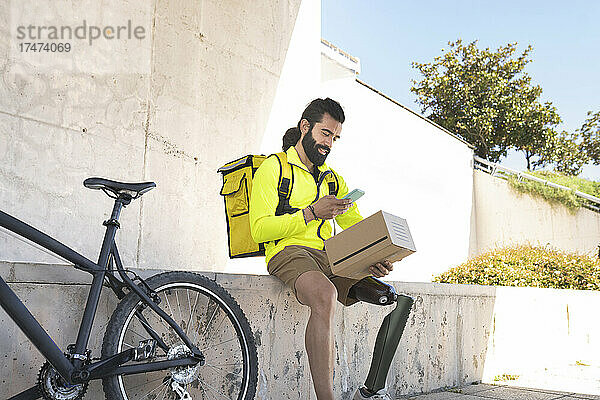 Lächelnder behinderter Lieferbote scannt Box mit Mobiltelefon mit dem Fahrrad