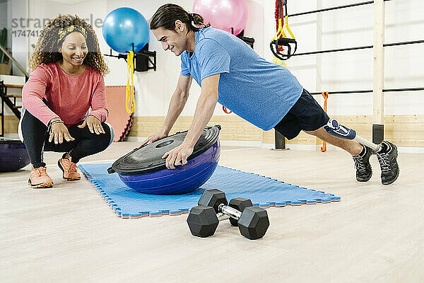 Lächelnde Trainerin unterstützt behinderten Mann bei Liegestützen auf dem Balanceball im Fitnessstudio