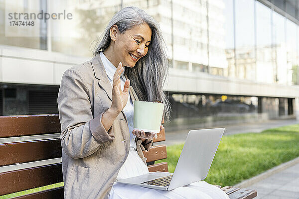 Frau grüßt während Videoanruf über Laptop  während sie auf Bank sitzt