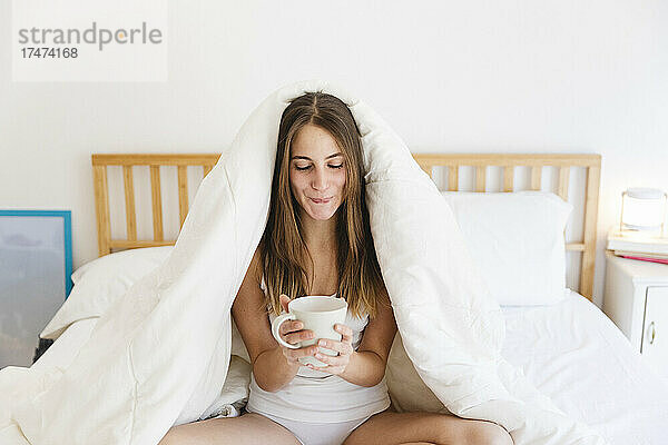 Junge Frau mit braunem Haar hält eine Tasse Kaffee auf dem Bett