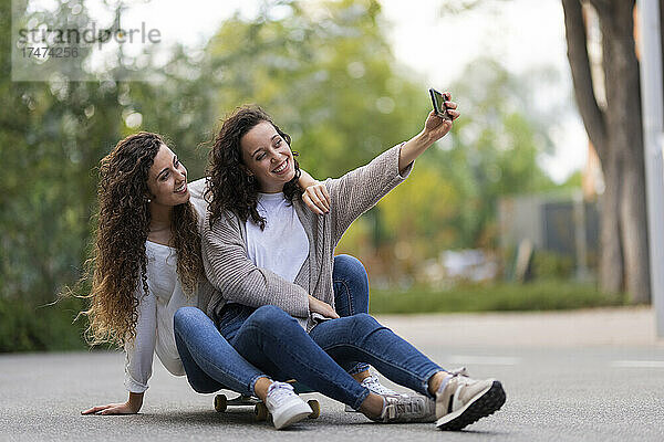 Lächelnde Frau macht ein Selfie mit ihrer Freundin per Smartphone  während sie auf dem Skateboard sitzt