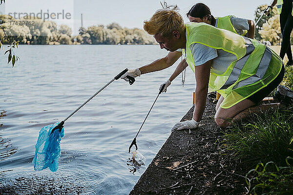 Männliche und weibliche Umweltschützer sammeln mit ihren Klauen Plastik aus dem See auf