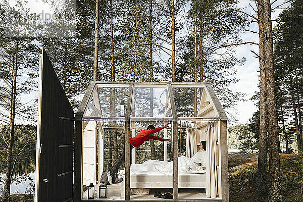 Mann springt  während seine Freundin in einem Glashaus auf dem Bett liegt