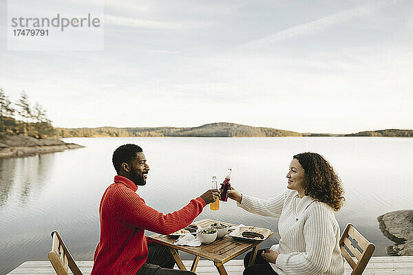 Lächelndes Paar stößt auf Flaschen an  während es am Wochenende auf einem Stuhl am See sitzt
