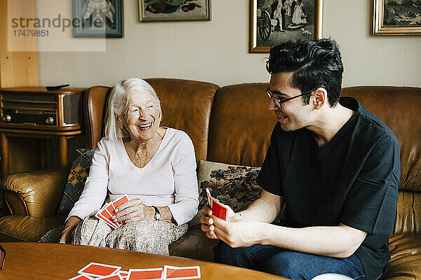 Glückliche männliche Pflegekraft  die mit einer älteren Frau im Wohnzimmer Karten spielt