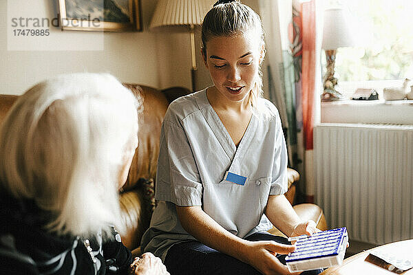 Weibliche Krankenschwester mit Medikamentenbox im Gespräch mit älterer Frau im Wohnzimmer
