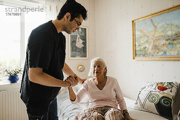 Männlicher Pfleger hält die Hand  während er eine ältere Frau unterstützt  die zu Hause auf dem Bett sitzt
