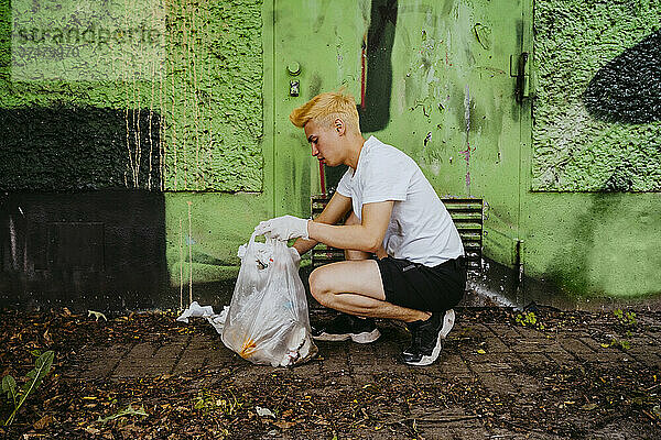 Seitenansicht eines jungen männlichen Freiwilligen in der Hocke beim Aufsammeln von Plastikmüll an einer grünen Wand