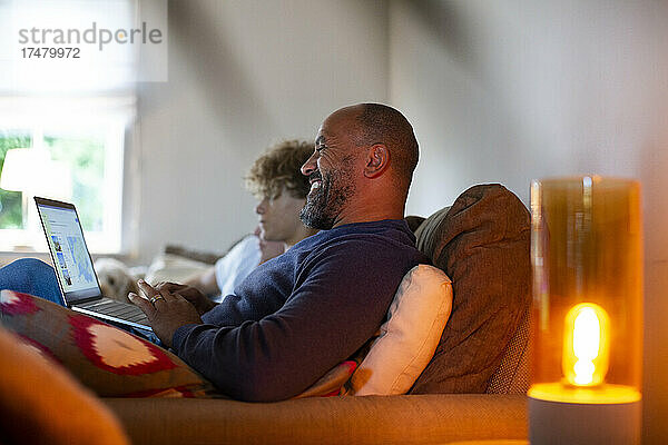 Lächelnder reifer Mann  der einen Laptop benutzt und neben seinem Sohn auf dem Sofa im Wohnzimmer sitzt