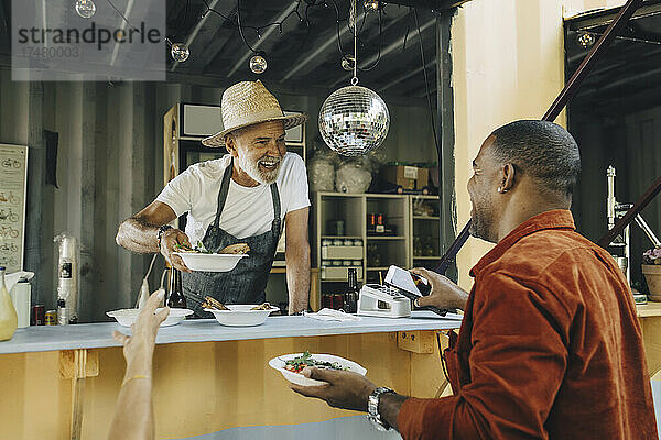 Lächelnder Besitzer  der Essen serviert  während ein männlicher Kunde mit seinem Smartphone kontaktlos bezahlt