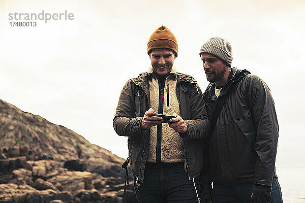 Lächelnder Mann teilt Smartphone mit männlichem Freund gegen den Himmel