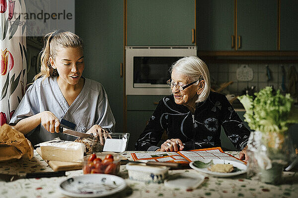 Weibliche Pflegekraft schneidet Brotlaib am Esstisch  während sie neben einer älteren Frau in der Küche sitzt