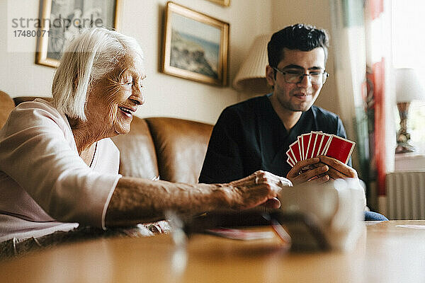 Glückliche ältere Frau spielt Karten mit einem männlichen Angestellten im Gesundheitswesen im Wohnzimmer