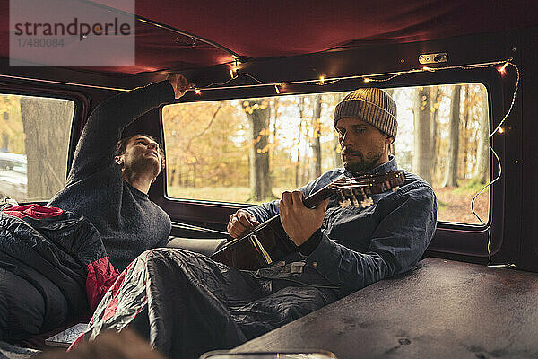 Mann spielt Gitarre  während sein Freund die Lichter im Campingbus betrachtet