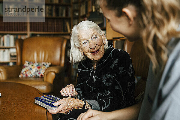 Lächelnde ältere Frau mit Medikamentenschachtel im Gespräch mit einer Mitarbeiterin des Gesundheitswesens im heimischen Wohnzimmer