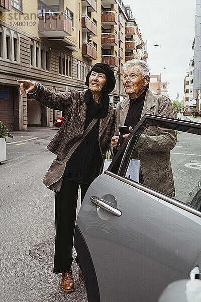 Lächelnde Frau  die auf ein Auto auf der Straße zeigt  während ein älterer Mann sein Smartphone in der Hand hält
