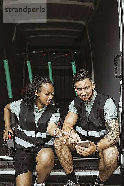 Lächelnde weibliche und männliche Mitarbeiter  die ihr Smartphone benutzen  während sie im Lieferwagen sitzen