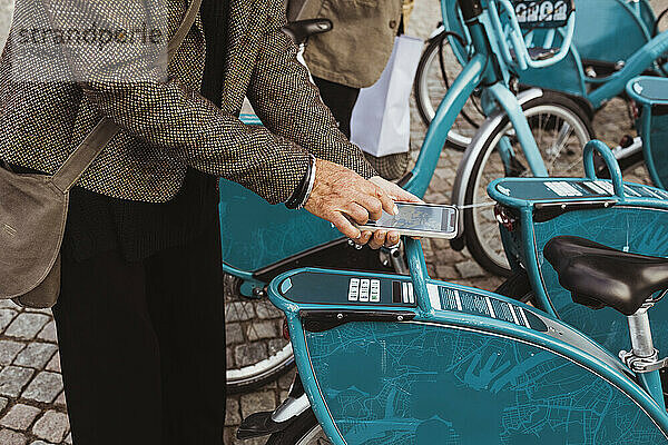 Mittelteil einer älteren Frau  die den QR-Code eines Fahrrads mit ihrem Smartphone an einer Parkstation scannt
