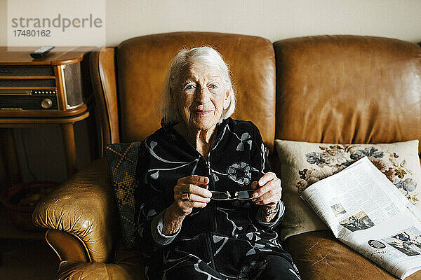 Porträt einer älteren Frau  die eine Brille hält und auf einem Sofa sitzt