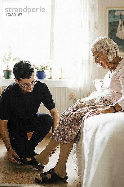 Lächelnde ältere Frau schaut  während männliche Krankenschwester ihr hilft  Sandale im Schlafzimmer zu setzen