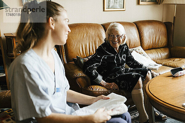Lächelnde ältere Frau im Gespräch mit weiblicher Gesundheitspflege  während sie im Wohnzimmer zu Hause sitzt