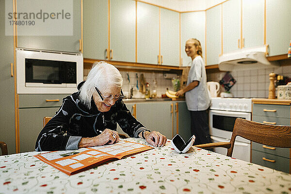 Lächelnde ältere Frau  die ein Sudoku in einem Buch löst  während eine weibliche Betreuerin zu Hause Küchenarbeiten erledigt