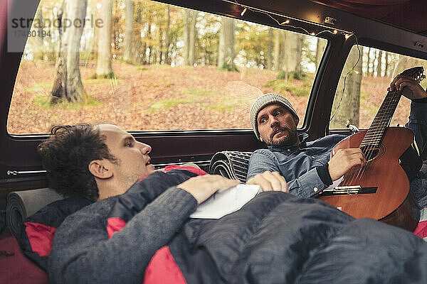 Mann zupft Gitarre  während er neben einem männlichen Freund im Campingwagen liegt