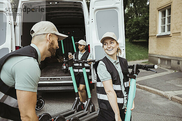 Lächelnde Zustellerin  die einen männlichen Mitarbeiter anschaut  während sie einen Motorroller in einen Lieferwagen lädt